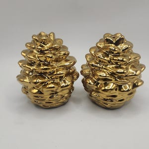 May include: Two gold-colored, pine cone-shaped decorative objects. The objects have a textured surface and are displayed on a white background. The objects are likely used for home decor.