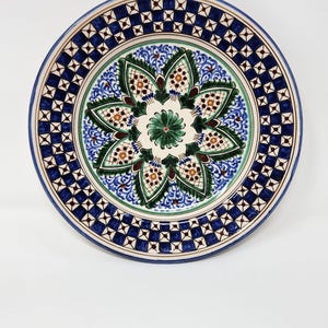 May include: A round ceramic plate with a detailed floral design. The plate features a blue border with a geometric pattern, and a central design with green, blue, and brown floral elements. The plate is set against a white background.