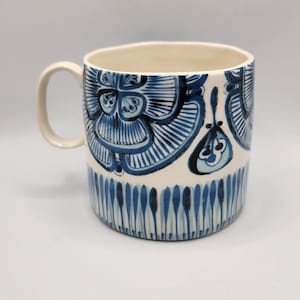 May include: A ceramic mug with a cream-colored interior and handle. The exterior is decorated with a blue floral pattern and vertical blue lines near the base. The mug is approximately 4 inches tall.