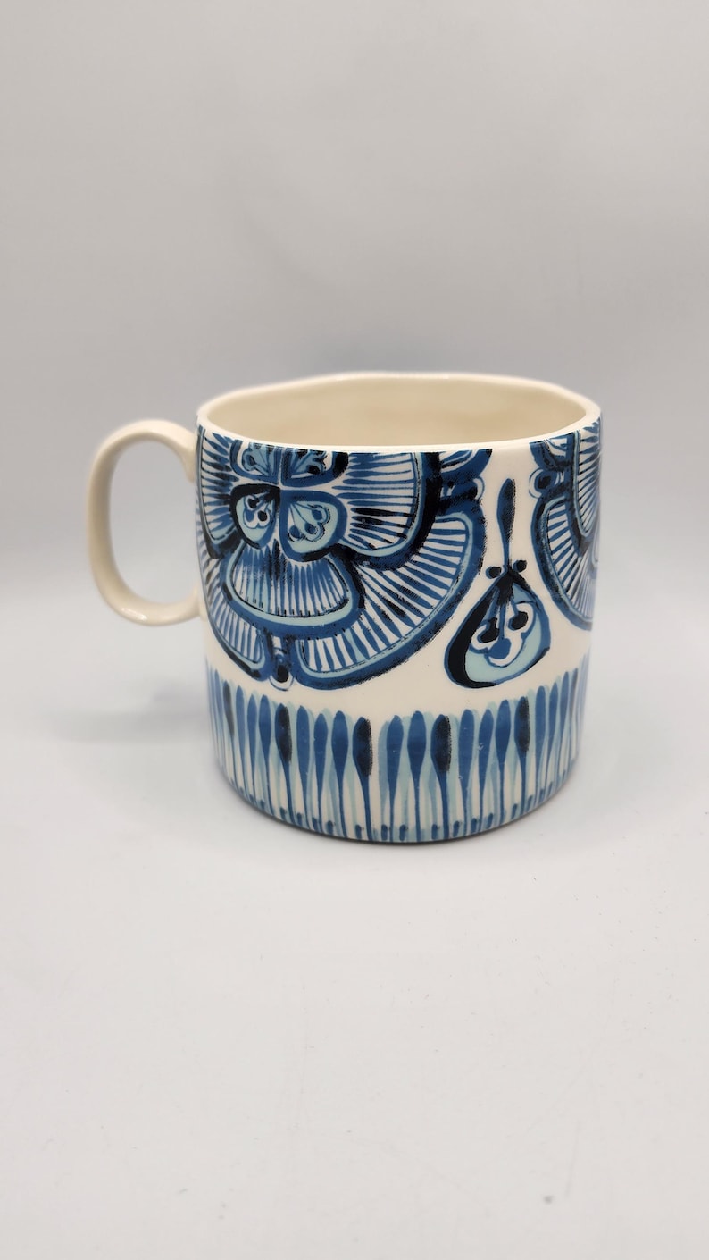 May include: A ceramic mug with a cream-colored interior and handle. The exterior is decorated with a blue floral pattern and vertical blue lines near the base. The mug is approximately 4 inches tall.