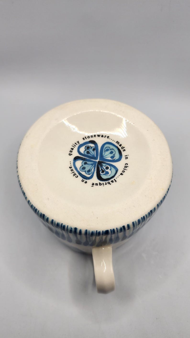 May include: White stoneware mug with a blue floral design and a handle. The mug has a blue rim and the words "quality stoneware...made in china...fabriqu&eacute; en chine" printed on the bottom.