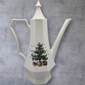 May include: White ceramic coffee pot with a Christmas tree design. The pot has a tall, angular body, a long spout, and a decorative handle. The Christmas tree is decorated with ornaments and presents.