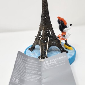 Pu&ograve; includere: Una miniatura della Torre Eiffel in bronzo, abbinata a una figura di Minnie Mouse con un vestito a pois e un fiocco. Le figure sono su una base blu. Un'etichetta con testo &egrave; di fronte.