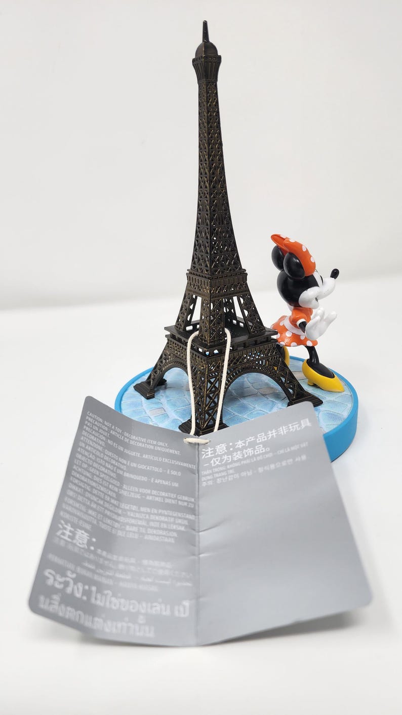 Pu&ograve; includere: Una miniatura della Torre Eiffel in bronzo, abbinata a una figura di Minnie Mouse con un vestito a pois e un fiocco. Le figure sono su una base blu. Un'etichetta con testo &egrave; di fronte.