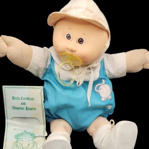 May include: A vintage Cabbage Patch Kids doll wearing a light blue onesie with an elephant applique, a white hat, and white shoes. The doll has a pacifier and comes with a birth certificate and adoption papers.