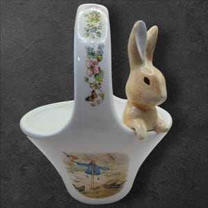 Può includere: Vaso da fiori in ceramica bianca a forma di cesto con una statuetta di coniglio sul lato. Il vaso ha un motivo floreale sul manico e un'illustrazione di Peter Rabbit sul fondo.