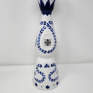 May include: A white ceramic bottle with blue floral designs and a silver cap. The bottle has a bulbous shape with a narrow neck and is decorated with blue leaf patterns and a central floral emblem.