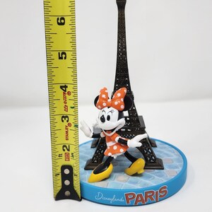 Pu&ograve; includere: Una statuina di Disneyland Paris con Minnie Mouse in un vestito e fiocco a pois arancioni, in piedi davanti a una Torre Eiffel in miniatura. La base &egrave; blu con la scritta "Disneyland Paris". La statuina &egrave; alta circa 7,6 cm.