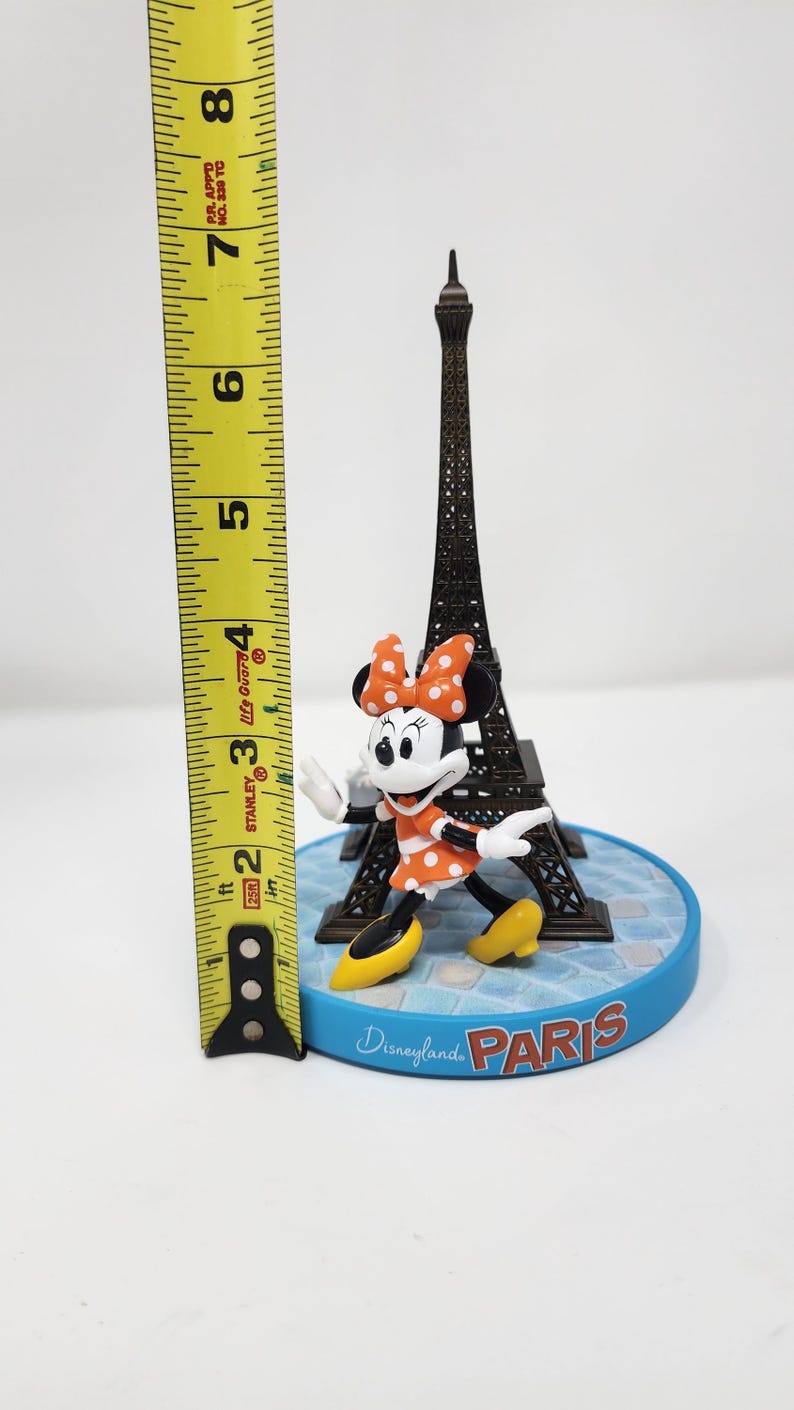 Pu&ograve; includere: Una statuina di Disneyland Paris con Minnie Mouse in un vestito e fiocco a pois arancioni, in piedi davanti a una Torre Eiffel in miniatura. La base &egrave; blu con la scritta "Disneyland Paris". La statuina &egrave; alta circa 7,6 cm.