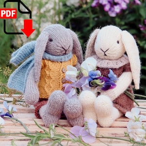 Puede incluir: Dos conejos de punto, uno gris con una bufanda azul y un suéter amarillo, el otro blanco con un vestido marrón, sosteniendo un ramo de flores moradas y blancas.
