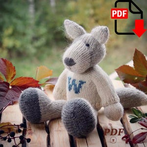 May include: A hand-knitted stuffed animal resembling a wolf or fox, featuring a light gray head with darker gray ears and snout, and a cream-colored body with a blue letter W embroidered on the chest. The toy has dark gray paws and a long tail.