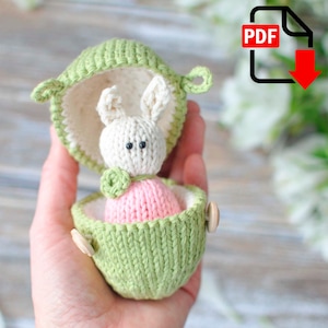 May include: A knitted bunny toy in a green pod. The bunny is white with black eyes and a pink body. The pod is light green with a white interior and wooden button closures. The image includes a PDF download icon.