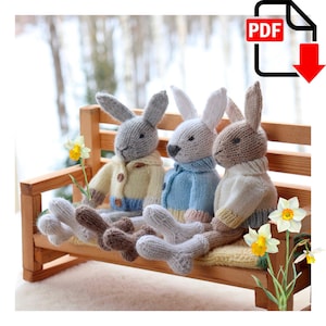 Pode incluir: Três coelhos de malha sentados em um banco de madeira. Os coelhos estão vestindo suéteres de diferentes cores: amarelo, azul e branco. Os coelhos estão sentados em um banco de madeira com um fundo branco.