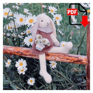 Puede incluir: Una muñeca de conejo tejida, con orejas largas, vestida con un vestido malva y sosteniendo una pequeña cesta de flores, está sentada en una valla de madera. El fondo presenta margaritas blancas y follaje verde. Un icono PDF con una flecha roja está en la esquina superior derecha.
