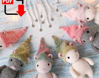 May include: A set of six knitted mouse ornaments with different coloured hats. The hats are pink, green, yellow, and grey. The mice are all wearing a grey knitted body and have a white face. The ornaments are sitting on a white doily on a wooden surface.