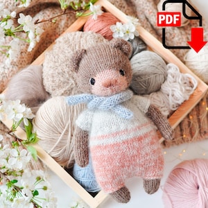 May include: A knitted brown bear toy with a light blue scarf, wearing a white and pink striped shirt. The bear is surrounded by balls of yarn in various colors, including beige, light blue, and pink.