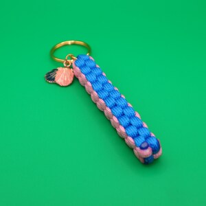 May include: A keychain featuring a rectangular braided paracord fob in bright blue and light pink. The fob is attached to a gold-toned metal ring, which also holds a small, dangling charm shaped like two layered seashells in pink and dark blue.