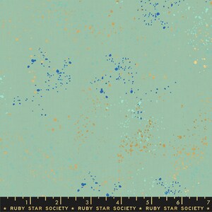 May include: A light green fabric with a speckled pattern of blue, gold, and light green dots. The fabric has a ruler printed at the bottom with the text "RUBY STAR SOCIETY" repeated.