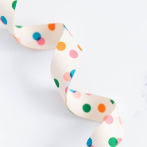 May include: A cream-coloured ribbon with a repeating pattern of colourful polka dots in shades of blue, green, orange, and pink. The ribbon is curled and appears to be made of fabric.