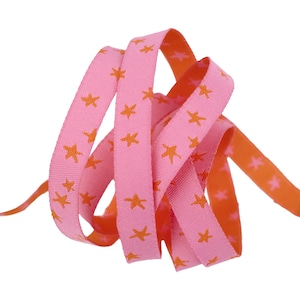 May include: Pink ribbon with orange star patterns. The ribbon is curled and layered, showcasing the design on both sides. The background is white, highlighting the ribbon's colors and patterns. This ribbon is suitable for crafts and gift wrapping.