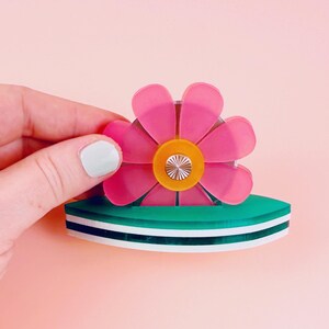 May include: A pink flower brooch with a yellow centre and a green base. The brooch has a layered design with white and black stripes. The flower petals are translucent.