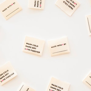 May include: A set of beige fabric labels with black text and a pink heart. The labels read "Wash Cold, Hang Dry, Love Forever" and "Made With Love By".