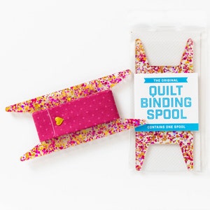 May include: A clear acrylic quilt binding spool with a pink fabric centre and gold heart accent. The spool is decorated with colourful glitter. The packaging reads "The Original Quilt Binding Spool".