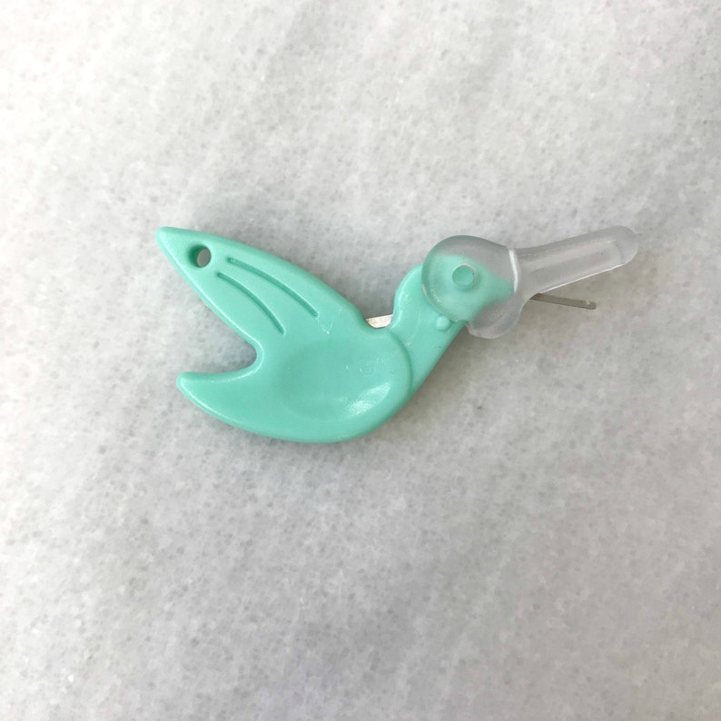 May include: A turquoise plastic bird-shaped threader with a clear plastic needle. The threader has a small hole on the top of the bird's head.