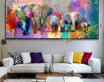 Puede incluir: Pintura abstracta de una familia de elefantes en un paisaje colorido. La obra presenta una paleta de colores vibrantes, incluyendo rojo, morado, azul y verde, con los elefantes en tonos grises. El cuadro está enmarcado en negro y cuelga sobre un sofá blanco.