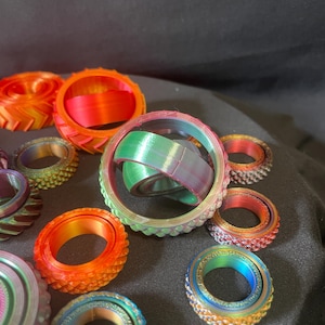 May include: A collection of colorful, 3D-printed rings in various sizes and designs. The rings feature a range of colors, including orange, red, green, blue, and purple, with textured surfaces and intricate patterns. Some rings have a gear-like appearance.
