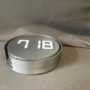 May include: A set of black coasters with the numbers "7 18" in white. The coasters are round and sit in a matching holder. The set is made of a matte material and is suitable for home or office use.