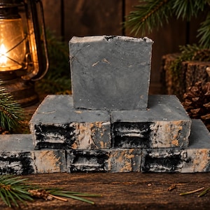 May include: A stack of five rectangular bars of charcoal-colored soap. The soap bars feature a smooth, dark gray top surface and a textured, layered side profile with visible streaks of black and tan colors throughout the soap's composition.