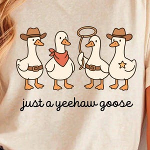 May include: A beige t-shirt featuring a cartoon design of four geese dressed as cowboys. The geese wear cowboy hats, bandanas, and belts. One goose holds a lasso. The text "just a yeehaw goose" is written in a script font below the geese.