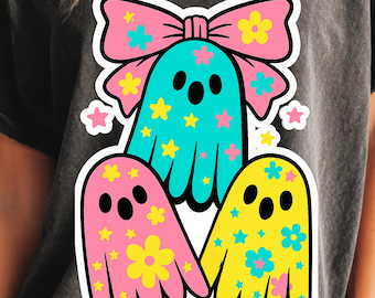 Può includere: Un design grafico con tre fantasmi dei cartoni animati rosa, blu e gialli, decorati con fiori e stelle. Un fiocco rosa con dettagli floreali è sopra il fantasma centrale. Il design è stampato su una maglietta grigio scuro.