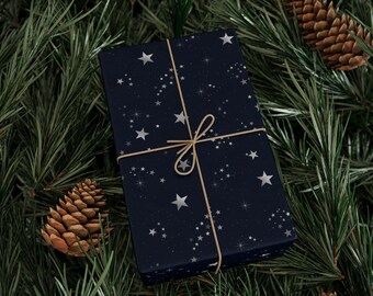 May include: A wrapped gift box with a dark blue background and a pattern of white stars and constellations. The box is tied with twine and rests on a bed of green pine needles and pine cones, creating a festive holiday scene.