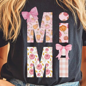 May include: Black t-shirt with the word "MIMI" in a fall-themed design. The letters are filled with pumpkins and floral patterns in pink and orange. Pink bows accent the letters, creating a festive autumn look.