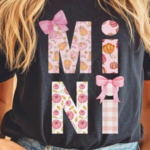 May include: Black t-shirt with the word "MINI" in a fall-themed design. The letters are filled with pumpkin and floral patterns, and accented with pink bows. The "I" is a vertical rectangle with pumpkins.