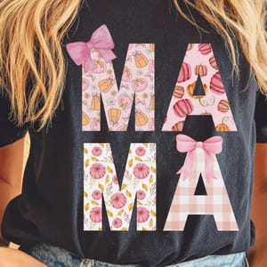 May include: Black t-shirt with the word "MAMA" in large, decorative letters. The letters are filled with patterns of pumpkins, flowers, and plaid, and adorned with pink bows. A fall-themed graphic tee.