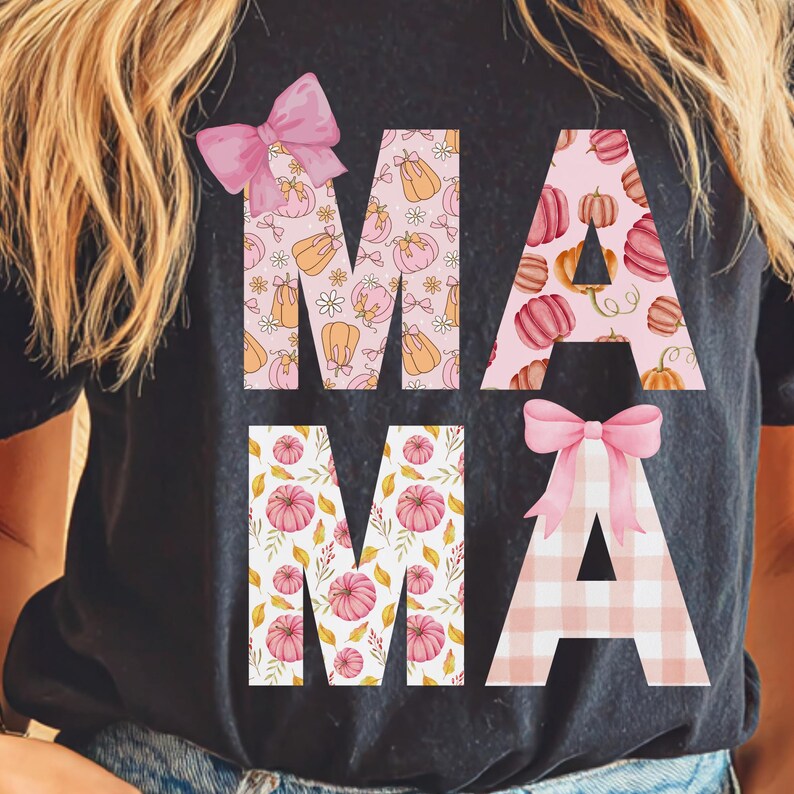 May include: Black t-shirt with the word "MAMA" in large, decorative letters. The letters are filled with patterns of pumpkins, flowers, and plaid, and adorned with pink bows. A fall-themed graphic tee.
