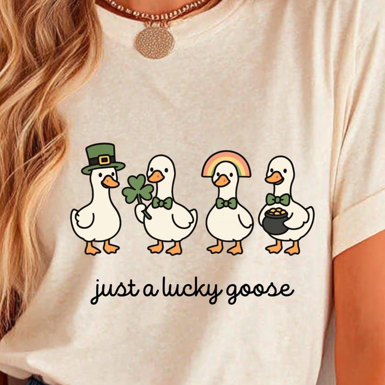 May include: Cream-colored t-shirt featuring a whimsical illustration of four cartoon geese. Each goose is adorned with St. Patrick's Day themed accessories, including a green hat, shamrock, rainbow, and pot of gold. The text "just a lucky goose" is printed below.