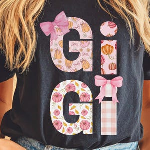 May include: Black t-shirt with the name "GiGi" in a fall-themed design. The letters are decorated with pumpkins, bows, and floral patterns in shades of pink, orange, and white.