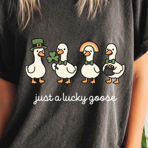 May include: Dark gray t-shirt featuring four cartoon geese with St. Patrick's Day themed accessories. One goose wears a green hat, another holds a shamrock, a third has a rainbow, and the last holds a pot of gold. The text "just a lucky goose" is below.