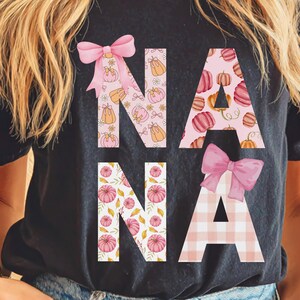 May include: Black t-shirt with the word "NANA" in pink and orange fall-themed patterns. The letters are decorated with pumpkins, bows, and floral accents, perfect for autumn. A stylish and festive design.