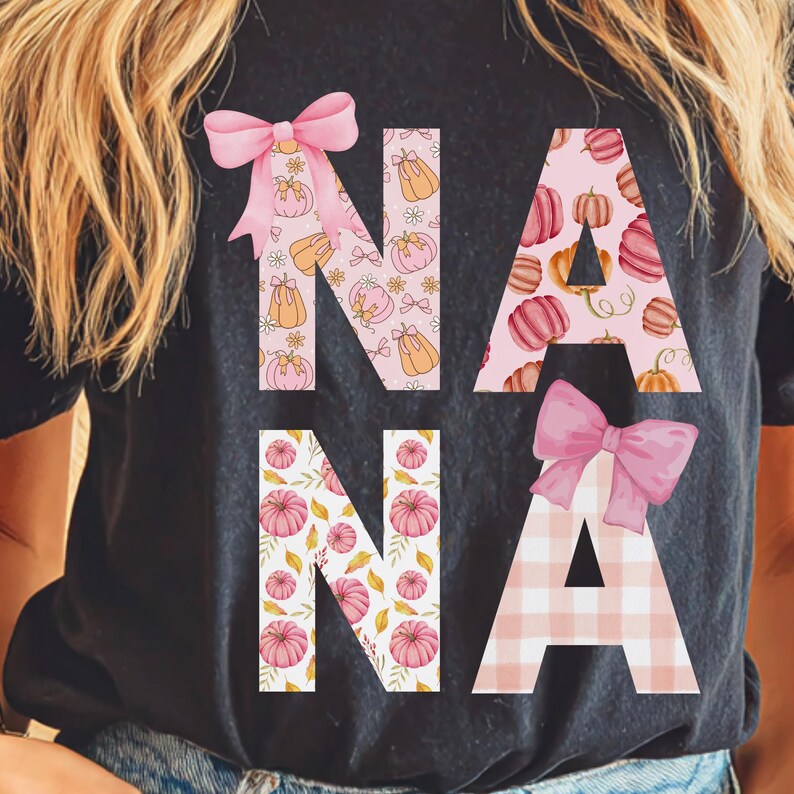 May include: Black t-shirt with the word "NANA" in pink and orange fall-themed patterns. The letters are decorated with pumpkins, bows, and floral accents, perfect for autumn. A stylish and festive design.