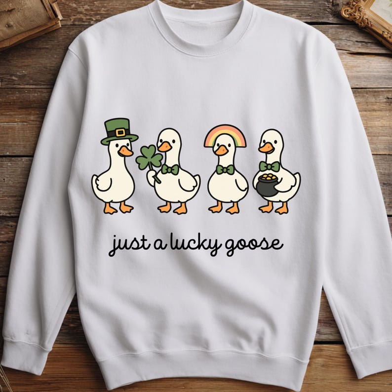May include: White sweatshirt featuring a cartoon design of four geese dressed for St. Patrick's Day. Each goose has a different accessory, including a hat, shamrock, rainbow, and pot of gold. The text "just a lucky goose" is printed below the geese.