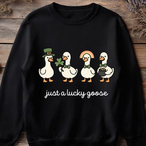 May include: Black sweatshirt featuring four cartoon geese, each adorned with St. Patrick's Day themed accessories: a green hat, a shamrock, a rainbow, and a pot of gold. The text "just a lucky goose" is written in cursive below the geese.