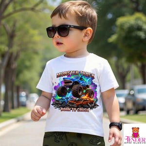 May include: White t-shirt with a colorful monster truck graphic. The design features a blue monster truck with a shark-like mouth, lightning bolts, and the text "MONSTER TRUCK" and "BORN TO CRUSH". The shirt is worn by a young child wearing sunglasses.