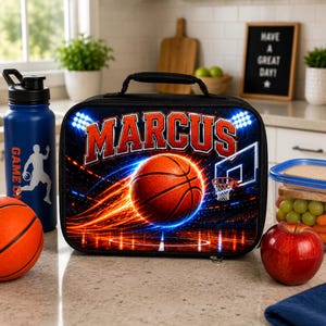 Könnte beinhalten: Eine rechteckige schwarze Lunchtasche mit einem Basketball-Motiv und dem Namen MARCUS in roten Buchstaben. Das Design zeigt ein Basketballfeld, einen Korb und leuchtende Lichtspuren. Die Tasche hat einen Tragegriff und einen Reißverschluss.