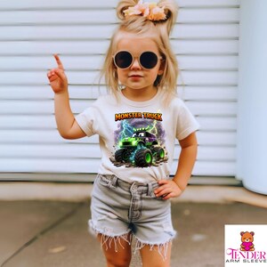 May include: A child wearing a cream-colored t-shirt with a graphic of a green and purple monster truck with lightning bolts and the words "MONSTER TRUCK". The child is also wearing denim shorts and sunglasses.