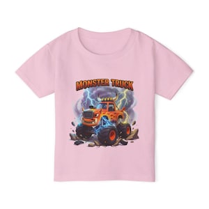 Puede incluir: Una camiseta rosa claro con un dise&ntilde;o de cami&oacute;n monstruo de dibujos animados. El cami&oacute;n naranja tiene neum&aacute;ticos negros grandes con llantas rojas, sobre un fondo de rel&aacute;mpagos y rocas. Las palabras "MONSTER TRUCK" est&aacute;n impresas encima del cami&oacute;n.